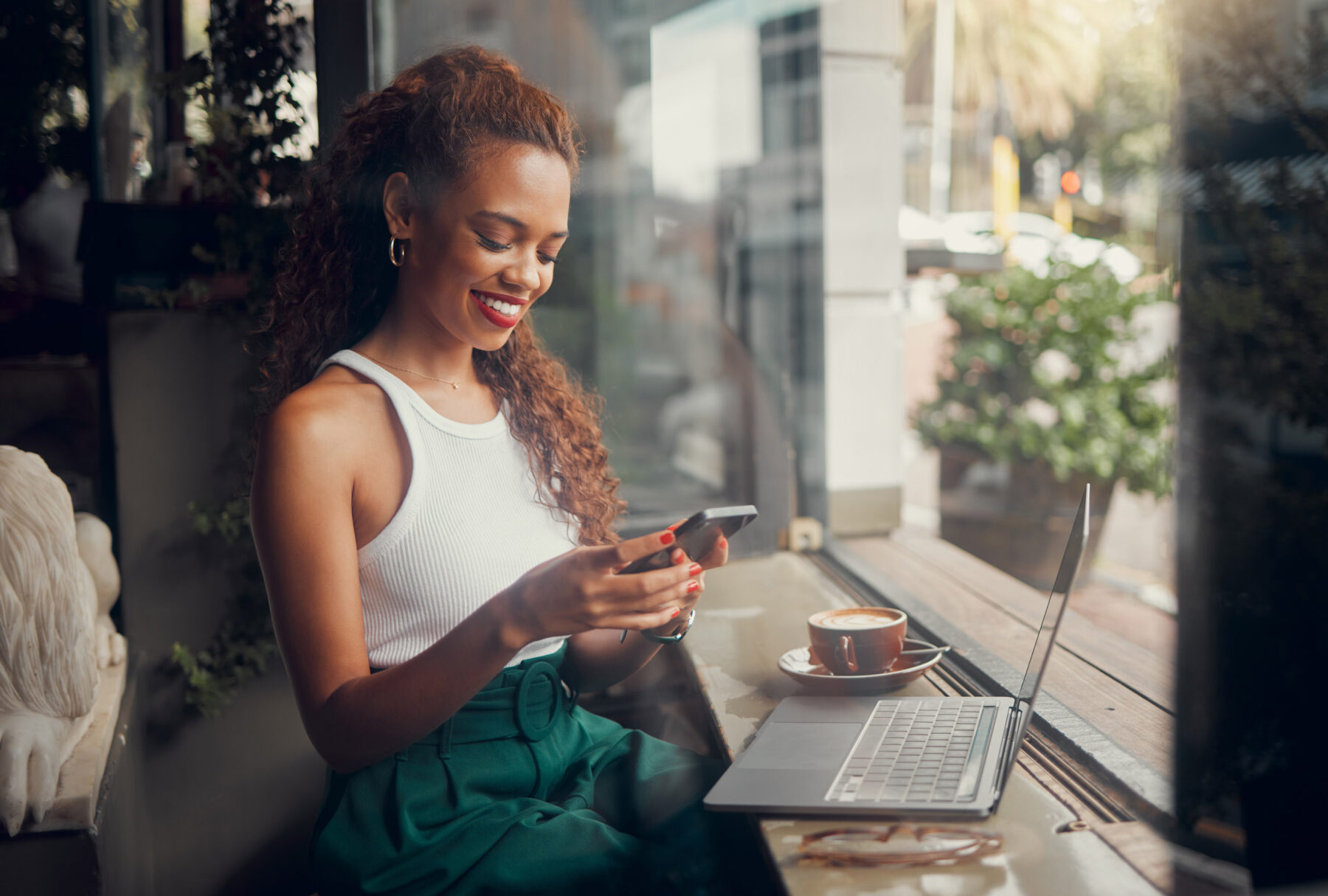 A woman sits by a window in a café, smiling at her smartphone. She has a laptop and a cup of coffee in front of her, and sunlight streams in, creating a warm and relaxed atmosphere.